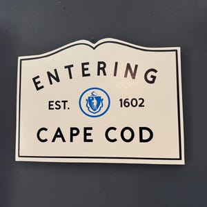 Peut inclure: Panneau rectangulaire blanc avec bordure noire et bord supérieur décoratif. Il est écrit "ENTERING CAPE COD" en noir, avec "EST. 1602" et un emblème bleu. Le panneau est fixé sur un mur bleu foncé.