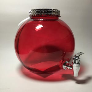May include: A spherical red glass beverage dispenser featuring a silver-colored metallic lid with a textured rim and a silver-colored spigot attached to the lower side. The transparent red glass body has a smooth, rounded shape and a wide opening.