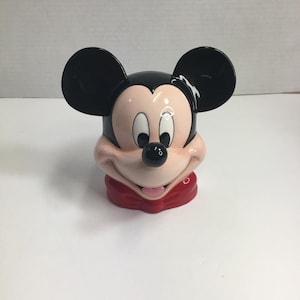 May include: A ceramic Mickey Mouse head-shaped container featuring large black ears, a pale peach-colored face, white eyes with black pupils, a small black nose, and a wide smiling mouth with a pink tongue, all resting on a red base.
