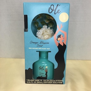 Puede incluir: Una caja azul claro para un difusor de varillas Olé Orange Blossom que contiene un jarrón de vidrio, cinco varillas de 22 cm y 90 ml de solución. El empaque presenta un gráfico de una bailarina y una ilustración de flor blanca.