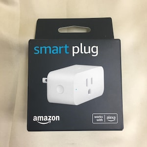 May include: A white Amazon smart plug in its black retail packaging. The box features the text smart plug at the top, the Amazon logo at the bottom left, and a works with alexa badge at the bottom right. The plug has a circular button on the side.