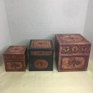Könnte beinhalten: Ein Set aus drei quadratischen Schachteln in abgestuften Größen mit einem abgenutzten braun-schwarzen Finish, verziert mit gemalten Herzmotiven und dekorativen Randmustern auf den Deckeln und Seiten, angeordnet in einer Reihe von klein nach groß.