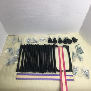 May include: A collection of matte black cabinet pulls and matching round knobs, accompanied by numerous small silver metal screws in plastic bags. A purple ruler measuring 12 inches and a pink ruler measuring 12 inches are placed underneath.