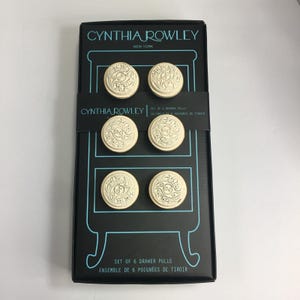 May include: A set of six round, cream-colored drawer pulls with intricate embossed floral patterns, presented in a black rectangular box labeled Cynthia Rowley New York. The packaging features a light blue outline of a dresser and text in English.