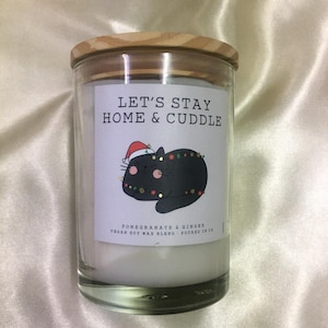 Könnte beinhalten: Ein Kerzenglas aus klarem Glas mit einem hellen Holzdeckel. Das weiße Etikett zeigt den Text LET'S STAY HOME & CUDDLE über einer schwarzen Katze mit Weihnachtsmütze und Lichterkette. Darunter steht POMEGRANATE & GINGER VEGAN SOY WAX BLEND.