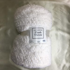 Pode incluir: Uma manta de pelúcia branca com um padrão texturizado em relevo, dobrada e presa com uma faixa de papel. A faixa apresenta o texto LIMITED EDITION Plush Throw, 127 cm x 152 cm, 100% poliéster, Made in China e o logotipo da marca ULTA.