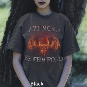Peut inclure: T-shirt noir avec le nom du groupe "AVENGED SEVENFOLD" en police texturée. Le t-shirt présente un motif de crâne enflammé. Le mot "Black" est imprimé en bas du t-shirt.