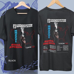 Op de afbeelding: Een zwart t-shirt met een grafische print van een hand die een diamant vasthoudt, een rode omtrek van de Verenigde Staten, de tekst "SOUNDSYSTEM", "MMXXVI", "NORTH AMERICA", "VICTORYLAND & FEIST" en een lijst met tourdata en locaties.