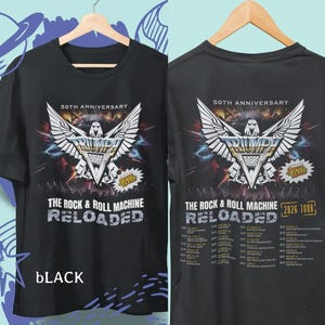 Könnte beinhalten: Schwarzes T-Shirt mit einem Grafikdesign, das das "50. JAHRESTAG" einer Band feiert, mit dem Text "THE ROCK & ROLL MACHINE RELOADED". Das Design beinhaltet ein geflügeltes Emblem und Tourdaten für 2026. Das Wort "BLACK" steht unten.