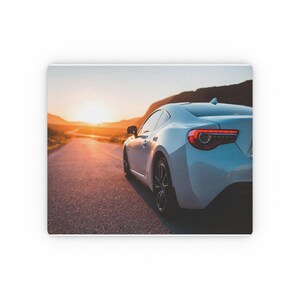 Op de afbeelding: Een witte sportwagen wordt van achteren gefotografeerd, rijdend op een weg bij zonsondergang. De achterlichten van de auto zijn rood en de wielen zijn zwart. De weg strekt zich uit in de verte, met bergen en een heldere zon op de achtergrond.