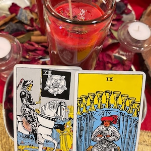 Pu&ograve; includere: Due carte dei tarocchi, Morte e Nove di Coppe, sono tenute davanti a una candela rossa in un barattolo di vetro. La carta della Morte raffigura uno scheletro a cavallo. La carta dei Nove di Coppe mostra una persona seduta davanti a nove coppe d'oro. Altre candele e petali di rosa sullo sfondo.