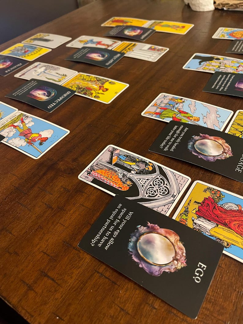 May include: A collection of tarot cards laid out on a dark wooden surface. The cards feature various illustrations and text, including the words "EGO" and "UNEXPECTED". Some cards have questions like "Will your ego allow space for us to have an equal partnership?"