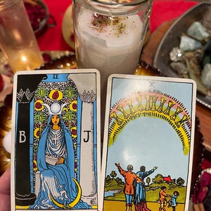 May include: Two tarot cards are held in front of a candle. The High Priestess card features a woman in blue robes. The other card shows a family under an arch of ten cups. A lit candle in a glass jar is in the background.