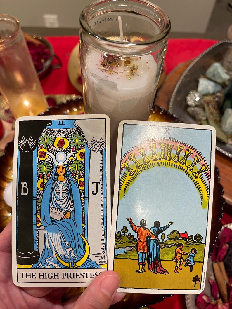 May include: Two tarot cards are held in front of a candle. The High Priestess card features a woman in blue robes. The other card shows a family under an arch of ten cups. A lit candle in a glass jar is in the background.