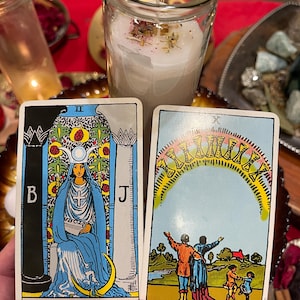 Pu&ograve; includere: Due carte dei tarocchi sono tenute davanti a una candela. La carta dell'Alta Sacerdotessa raffigura una donna in abito blu. L'altra carta mostra una famiglia sotto un arco di dieci coppe. Sullo sfondo c'&egrave; una candela accesa in un barattolo di vetro.