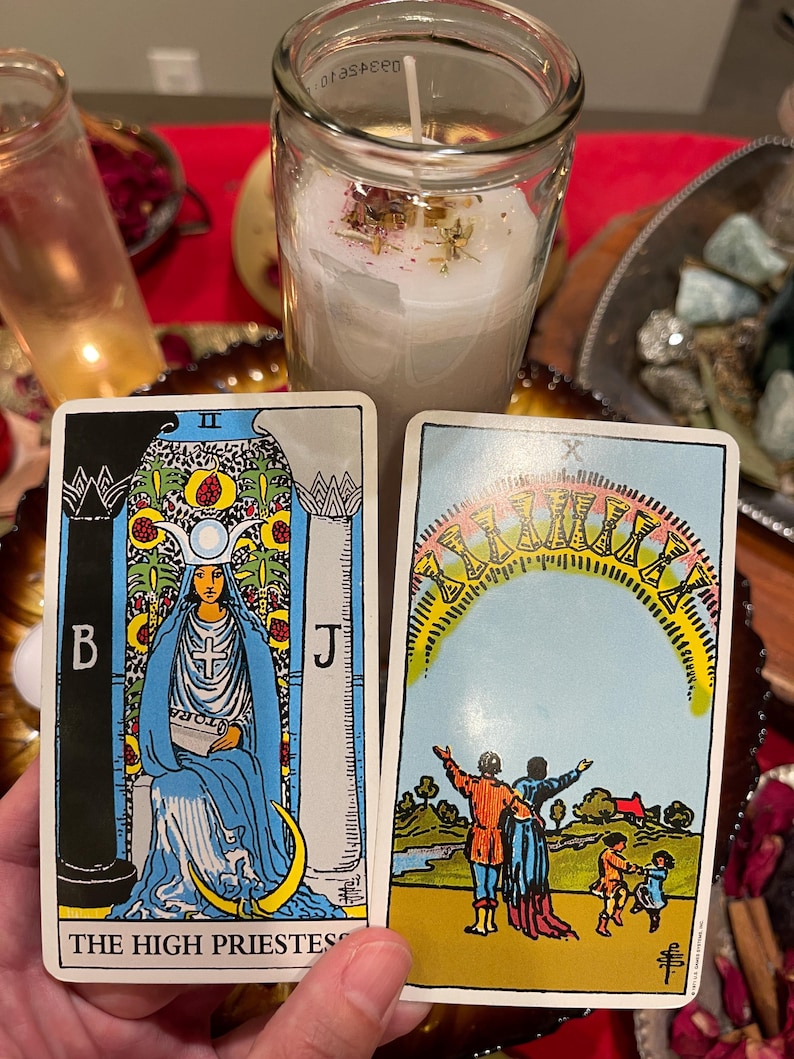 Pu&ograve; includere: Due carte dei tarocchi sono tenute davanti a una candela. La carta dell'Alta Sacerdotessa raffigura una donna in abito blu. L'altra carta mostra una famiglia sotto un arco di dieci coppe. Sullo sfondo c'&egrave; una candela accesa in un barattolo di vetro.