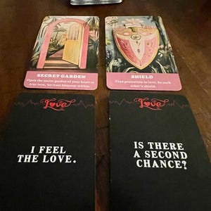 May include: A collection of tarot cards with various designs and text. Two cards feature illustrations of a garden and a shield. Two black cards read "I FEEL THE LOVE" and "IS THERE A SECOND CHANCE?" The cards are on a wooden surface.