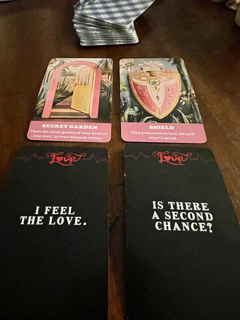 May include: A collection of tarot cards with various designs and text. Two cards feature illustrations of a garden and a shield. Two black cards read "I FEEL THE LOVE" and "IS THERE A SECOND CHANCE?" The cards are on a wooden surface.