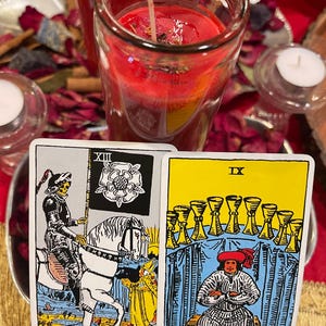May include: Two tarot cards are displayed, one depicting Death, the other IX of Cups. A red candle in a glass jar and two small white candles are in the background. Dried rose petals and other items are scattered around the candles.