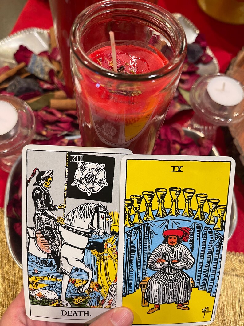 May include: Two tarot cards are displayed, one depicting Death, the other IX of Cups. A red candle in a glass jar and two small white candles are in the background. Dried rose petals and other items are scattered around the candles.
