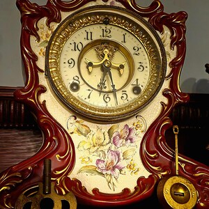 May include: Antique mantel clock with a red and gold frame. The clock face is cream-colored with gold numerals and hands. Floral designs in yellow and purple adorn the face. Includes a key and pendulum.
