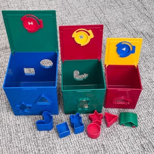 May include: Three colorful shape sorter boxes with matching shapes. The boxes are blue, green, and red, with yellow lids. The shapes are blue, red, and green. The shapes include a star, a triangle, a circle, and a square. Educational toy.