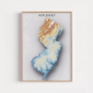 May include: A framed print featuring a 3D topographic map of New Jersey. The map is rendered in shades of blue, tan, and brown, with the state's name printed in black at the top. The artwork is set against a light gray background.