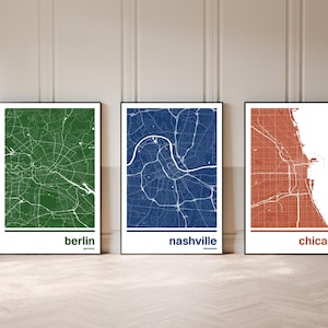 Puede incluir: Tres impresiones enmarcadas de mapas de ciudades. Los mapas son en verde, azul y naranja, con los nombres de las ciudades Berlín, Nashville y Chicago impresos debajo. Los mapas se muestran en una pared y un suelo de color claro.