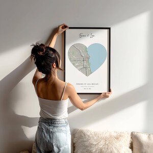 May include: A framed print with a heart-shaped map of Chicago, Illinois, and a light blue lake. The text reads "Emma & Ian" and "WHERE IT ALL BEGAN". The frame is black. The print is being held up against a white wall.