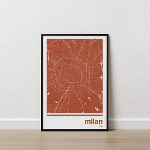 Puede incluir: Impresión enmarcada de un mapa de Milán, Italia, en color naranja quemado con líneas blancas que representan las calles. El nombre de la ciudad, "milan", está impreso en negrita en la parte inferior, con "Italy" debajo. El marco es negro.
