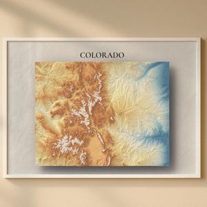May include: Framed topographic map of Colorado, featuring a detailed relief in shades of brown, tan, and blue. The word "COLORADO" is printed in a simple font above the map. The artwork is displayed in a white frame.