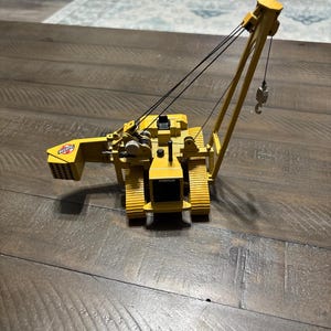 Pu&ograve; includere: Un modello di gru cingolata Caterpillar gialla con cingoli neri e un braccio a traliccio. La macchina presenta un logo Bechtel sul contrappeso posteriore, una cabina operatore nera e un gancio metallico sospeso da cavi neri.