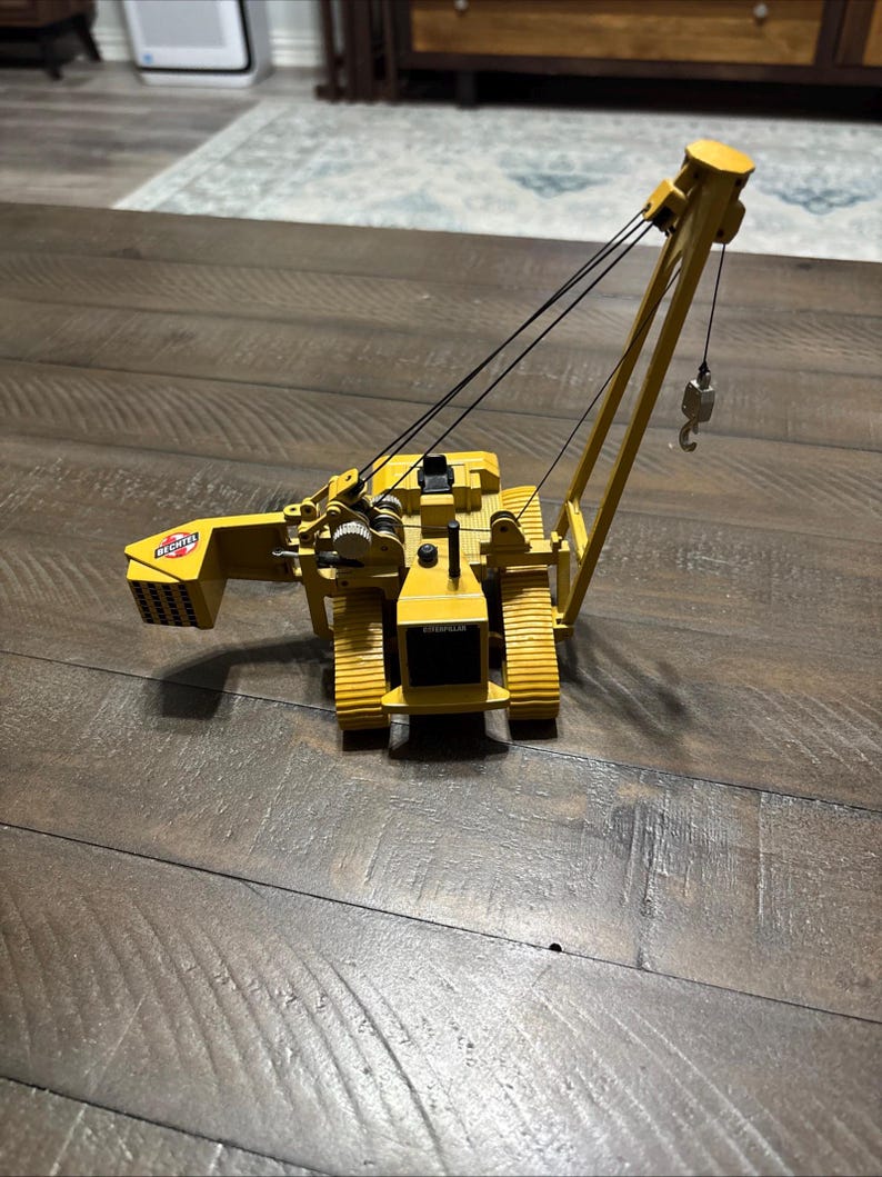Pu&ograve; includere: Un modello di gru cingolata Caterpillar gialla con cingoli neri e un braccio a traliccio. La macchina presenta un logo Bechtel sul contrappeso posteriore, una cabina operatore nera e un gancio metallico sospeso da cavi neri.