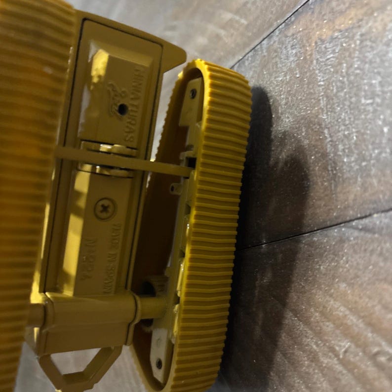 Pu&ograve; includere: Un gruppo cingoli per veicolo da costruzione giocattolo in plastica gialla con un motivo a battistrada scanalato. Il telaio laterale presenta il testo in rilievo MINIATURAS, MADE IN SPAIN e N&deg;824, insieme a una vite metallica di fissaggio.
