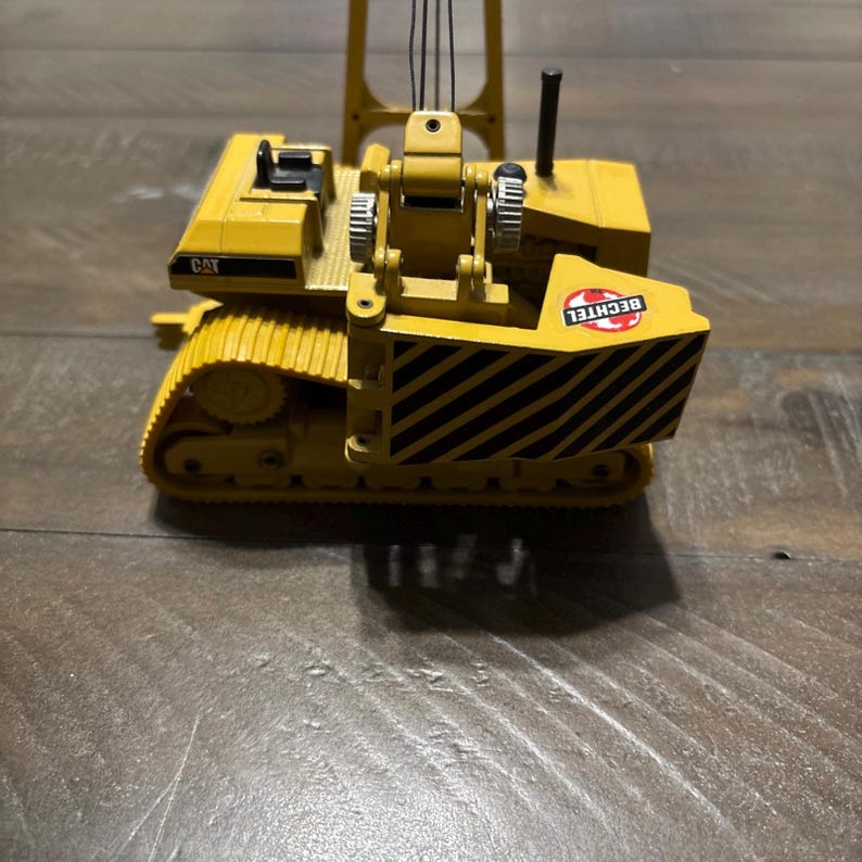 Pu&ograve; includere: Un modellino di gru cingolata Caterpillar gialla con cingoli neri, sedile dell'operatore nero e un contrappeso con strisce diagonali nere e gialle. Il logo Bechtel &egrave; stampato sul lato dell'unit&agrave; del contrappeso.