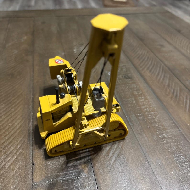 Pu&ograve; includere: Un modellino in metallo giallo di un veicolo da costruzione posatubi Caterpillar 591. Il modello presenta un braccio alto, un gancio in metallo, cavi neri e cingoli gialli. Il numero 591 e un logo sono visibili sul lato della cabina.
