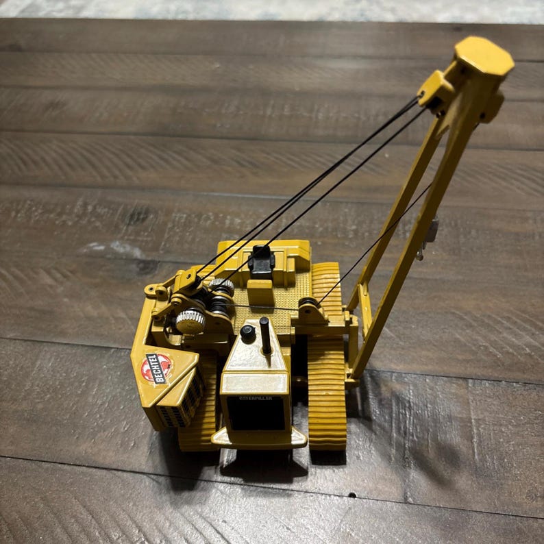 Pu&ograve; includere: Un modellino in scala di una gru cingolata Caterpillar gialla con sedile operatore nero, cingoli neri e braccio con cavi neri. Il lato della macchina presenta un logo Bechtel bianco con accenti rossi e il marchio Caterpillar.