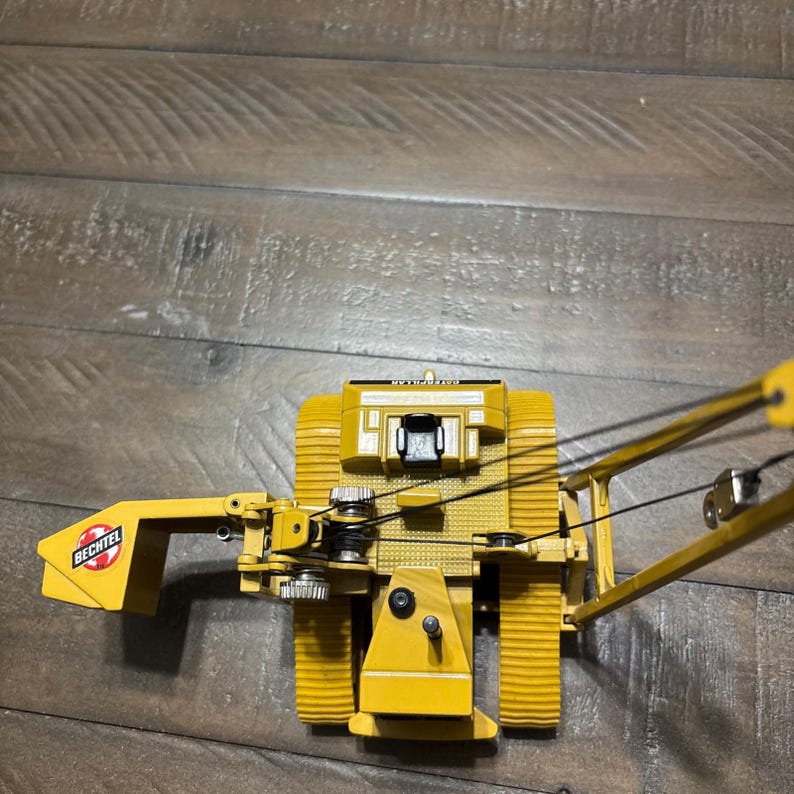 Pu&ograve; includere: Un modellino di veicolo da costruzione posatubi Caterpillar giallo con sedile operatore nero, braccio metallico con cavi neri e un contrappeso laterale chiaramente contrassegnato dal logo Bechtel rosso e bianco su una superficie gialla.