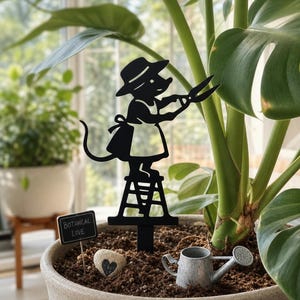 May include: A black metal plant stake featuring a silhouette of a mouse wearing a hat and apron while standing on a ladder and holding pruning shears. The stake is inserted into soil alongside a small metal watering can and a heart-shaped stone.