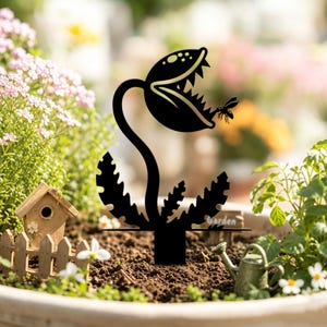 Può includere: Un picchetto da giardino in metallo nero raffigurante la sagoma di una pianta carnivora con una bocca spalancata, denti affilati e una piccola mosca vicino alla mascella. La pianta ha uno stelo curvo e foglie seghettate alla base.