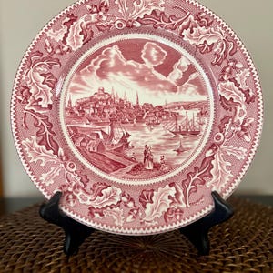 May include: A round, red and white decorative plate with a detailed illustration of a harbor scene. The plate features a scalloped edge and a border of leaves and acorns. The harbor scene includes buildings, boats, and people.