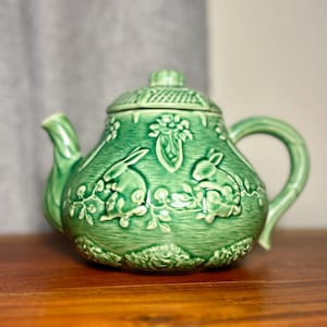 May include: Green ceramic teapot with a lid, featuring embossed designs of rabbits, flowers, and foliage. The teapot has a curved handle and a spout. The teapot is sitting on a wooden surface.