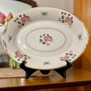 May include: An oval, off-white serving platter with a gold rim and floral bouquet designs. The platter features pink, blue, and yellow flowers. The center has a decorative dotted border. The platter is displayed on a black stand.