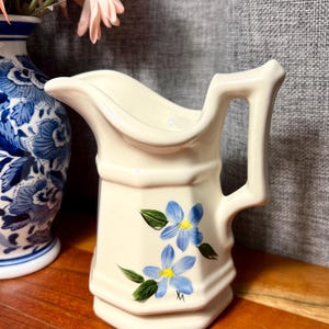May include: Cream-colored ceramic pitcher with a handle and a spout. The pitcher is decorated with two blue floral designs with green leaves. The pitcher has a hexagonal base and a decorative handle. A blue and white vase is partially visible in the background.