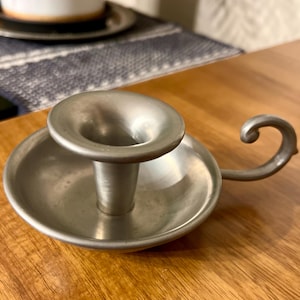 May include: A small, antique pewter candlestick holder with a circular base and a flared top. It features a handle with a curled end. The candlestick holder is displayed on a wooden surface.