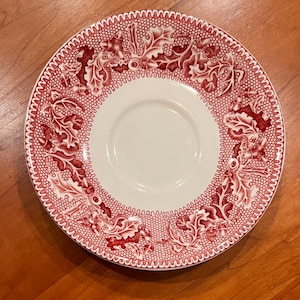 May include: A round ceramic saucer featuring a cream-colored center and a wide border decorated with a detailed red transferware pattern of oak leaves and acorns set against a fine, intricate mesh background texture.