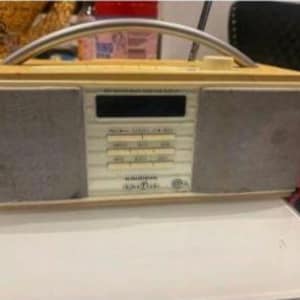 May include: A retro-style portable radio with a light yellow top and a silver handle. The sides are grey, and the front panel features a digital display and several buttons. The radio has the words "Audio Sonic" printed on the front.