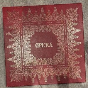 Op de afbeelding: Een vintage rode doos met gouden sierlijke details en het woord "OPERA" in het midden. De doos bevat de namen van componisten rond de rand, waaronder Verdi, Wagner en Rossini. De doos is waarschijnlijk voor een verzameling opera-opnames.