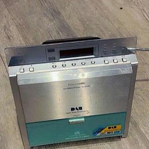 May include: A silver and teal Panasonic CD stereo system with DAB (Digital Audio Broadcasting) capabilities. It features a CD player, an LCD display, and control buttons. The unit is on a wooden surface.