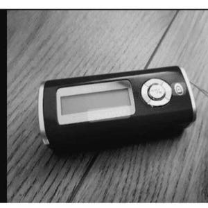 Puede incluir: Un reproductor de audio digital negro y plateado con una pantalla rectangular y botones de control. El dispositivo tiene un borde plateado. La pantalla está en blanco. El dispositivo está sobre una superficie de madera.